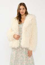 [Color: Vanilla] A front facing image of a brunette model wearing an ivory shaggy fur coat over a mint floral dress. 