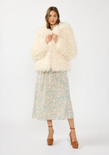 [Color: Vanilla] A front facing image of a brunette model wearing an ivory shaggy fur coat over a mint floral dress. 