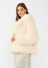 [Color: Vanilla] A side facing image of a brunette model wearing an ivory shaggy fur coat over a mint floral dress. 