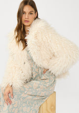 [Color: Vanilla] A sitting image of a brunette model wearing an ivory shaggy fur coat over a mint floral dress. 
