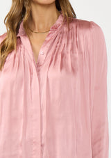[Color: Rose] A detail image of a brunette model wearing a pink silky poly satin blouse with pleats, a button front, and long sleeves. 