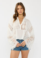 [Color: Off White] A front facing image of a brunette model wearing a white lace patchwork bohemian blouse with long bell sleeves and a v neckline. 