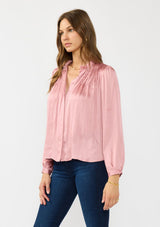 [Color: Rose] A side facing image of a brunette model wearing a pink silky poly satin blouse with pleats, a button front, and long sleeves. 