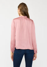 [Color: Rose] A back facing image of a brunette model wearing a pink silky poly satin blouse with pleats, a button front, and long sleeves. 