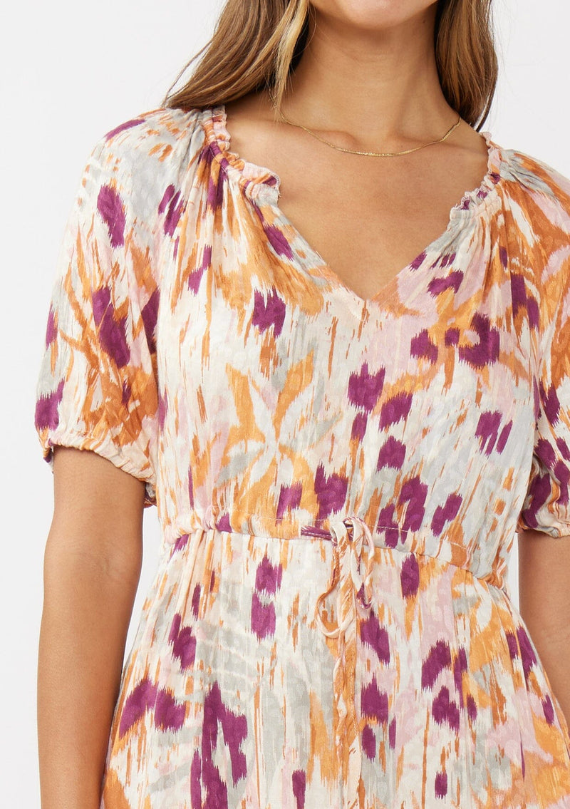 [Color: Natural/Plum] A detail image of a brunette model wearing an orange and purple midi dress with an abstract ikat print. A standout bohemian midi dress with short puff sleeves, split v neckline, drawstring waist, and a ruffle trimmed hem. A versatile midi dress styled with chucky clogs for the perfect boho chic look. 