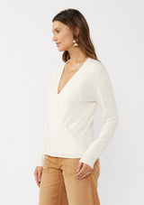 [Color: Off White] A side facing image of a brunette model wearing an off white solid knit sweater with a wrap front and side button closures. A soft versatile sweater, perfect for spring styling. 