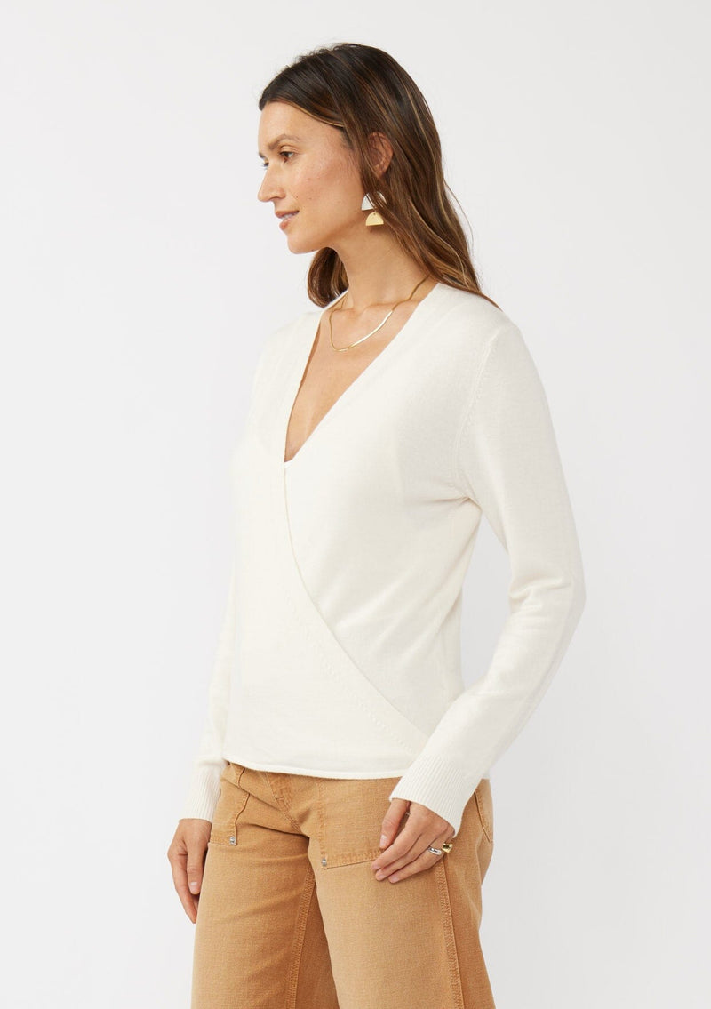[Color: Off White] A side facing image of a brunette model wearing an off white solid knit sweater with a wrap front and side button closures. A soft versatile sweater, perfect for spring styling. 