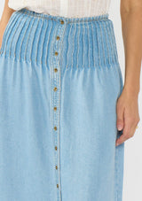 [Color: Light Wash] A detail image of a brunette model wearing a soft blue tencel maxi skirt with pintucks at the waist, a button front, and a smocked back. 