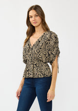[Color: Black/Natural] A side facing image of a brunette model wearing a black and beige paisley print top with ruched sleeves, a v neckline, and an elastic peplum waist. 