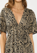 [Color: Black/Natural] A detail image of a brunette model wearing a black and beige paisley maxi dress with short sleeves, a deep v neckline, and a side slit. 