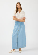 [Color: Light Wash] A front facing image of a brunette model wearing a white blouse with a soft blue tencel maxi skirt with pintucks at the waist, a button front, and a smocked back. 