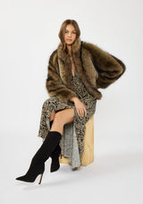 [Color: Black/Natural] A sitting image of a brunette model wearing a brown fur coat over a black and beige paisley maxi dress with short sleeves, a deep v neckline, and a side slit. 