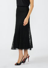 [Color: Black] A side facing image of a model wearing a black lace maxi skirt with black patent pumps. 