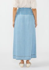 [Color: Light Wash] A back facing image of a brunette model wearing a white blouse with a soft blue tencel maxi skirt with pintucks at the waist, a button front, and a smocked back. 