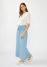 [Color: Light Wash] A side facing image of a brunette model wearing a white blouse with a soft blue tencel maxi skirt with pintucks at the waist, a button front, and a smocked back. 