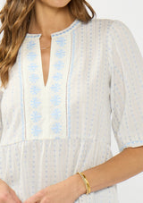 [Color: Light Blue/Cream] A detail image of a brunette model wearing a white and blue embroidered blouse with a peplum silhouette, split v neckline, and three quarter length sleeves.