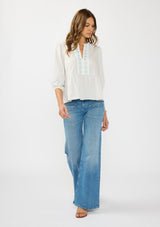 [Color: Light Blue/Cream] A front facing image of a brunette model wearing a white and blue embroidered blouse with a peplum silhouette, split v neckline, and three quarter length sleeves.