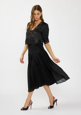 [Color: Black] A side facing image of a model wearing a black satin lace top with a crinkled black midi skirt.
