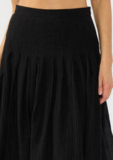 [Color: Black] A detail image of a model wearing a crinkled black midi skirt with pleats. Styled with black patent pumps.
