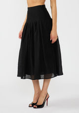 [Color: Black] A side facing image of a model wearing a crinkled black midi skirt with pleats. Styled with black patent pumps. 