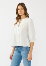 [Color: Light Blue/Cream] A side facing image of a brunette model wearing a white and blue embroidered blouse with a peplum silhouette, split v neckline, and three quarter length sleeves.