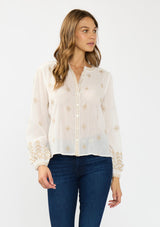 [Color: White/Sand] A front facing image of a brunette model wearing a white bohemian embroidered blouse with a button front. Styled with blue denim bottoms.