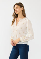 [Color: White/Sand] A side facing image of a brunette model wearing a white bohemian embroidered blouse with a button front. Styled with blue denim bottoms.