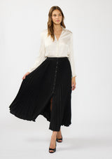 [Color: Black] A front facing image of a model wearing a white satin blouse with a black pleated maxi skirt with a button front and slit. 
