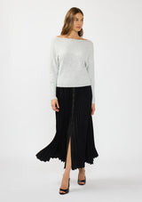 [Color: Heather Lunar] A front facing image of a brunette model wearing a light grey pullover sweater with an off shoulder design, boat neckline, and ribbed cuffs. Styled with a black pleated maxi skirt.