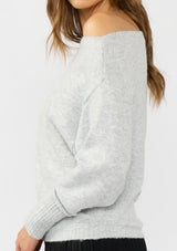 [Color: Heather Lunar] A  detail image of a brunette model wearing a light grey pullover sweater with an off shoulder design, boat neckline, and ribbed cuffs. 