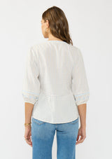 [Color: Light Blue/Cream] A back facing image of a brunette model wearing a white and blue embroidered blouse with a peplum silhouette, split v neckline, and three quarter length sleeves.
