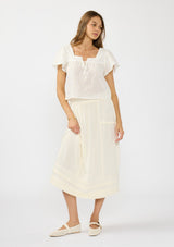 [Color: Natural] A front facing image of a brunette model wearing a white flutter sleeve top with an off white midi skirt with lace trims and pintucks. 