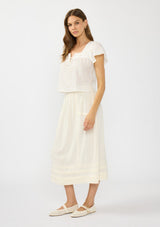 [Color: Natural] A side facing image of a brunette model wearing a white flutter sleeve top with an off white midi skirt with lace trims and pintucks. 