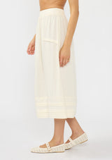 [Color: Natural] A side facing image of a model wearing an off white midi skirt with lace trims and pintucks. 