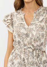 [Color: Cream/Black] A detail image of a brunette model wearing a cream and black floral mini dress with flutter sleeves, a split v neckline, lace trims, and a self tie waist belt. 