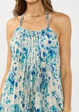 [Color: Natural/Aqua] A detail image of a brunette model wearing a flowy blue floral maxi dress with a pleated design, scoop neck, halter tie, and front slit. 