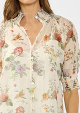 [Color: Ivory/Rosewood] A detail image of a brunette model wearing an ivory floral print top with puff sleeves, smocked cuffs, smocked neck, and a button front.
