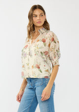 [Color: Ivory/Rosewood] A side facing image of a brunette model wearing an ivory floral print top with puff sleeves, smocked cuffs, smocked neck, and a button front.