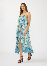 [Color: Natural/Aqua] A side facing image of a brunette model wearing a flowy blue floral maxi dress with a pleated design, scoop neck, halter tie, and front slit. 