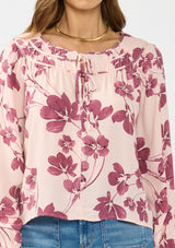 [Color: Coral/Wine] A detail image of a brunette model wearing a pink and wine floral blouse with a shirred yoke, double tie detail, and long sleeves with ruffled elastic cuffs. 