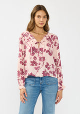 [Color: Coral/Wine] A front facing image of a brunette model wearing a pink and wine floral blouse with a shirred yoke, double tie detail, and long sleeves with ruffled elastic cuffs. 