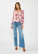 [Color: Coral/Wine] A front facing image of a brunette model wearing a pink and wine floral blouse with a shirred yoke, double tie detail, and long sleeves with ruffled elastic cuffs. 