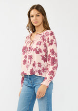[Color: Coral/Wine] A side facing image of a brunette model wearing a pink and wine floral blouse with a shirred yoke, double tie detail, and long sleeves with ruffled elastic cuffs. 