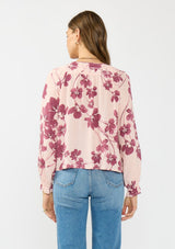[Color: Coral/Wine] A back facing image of a brunette model wearing a pink and wine floral blouse with a shirred yoke, double tie detail, and long sleeves with ruffled elastic cuffs. 