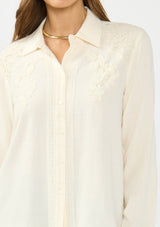 [Color: Natural] A detail image of a brunette model wearing an off white lace shirt with a collared neck and button front. 