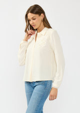 [Color: Natural] A side facing image of a brunette model wearing an off white lace shirt with a collared neck and button front. 