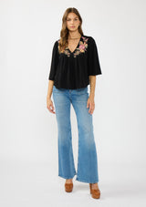 [Color: Black/Dusty Rose] A front facing image of a brunette model wearing a black embroidered top with flowy wide sleeves and a v neckline. 