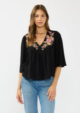 [Color: Black/Dusty Rose] A front facing image of a brunette model wearing a black embroidered top with flowy wide sleeves and a v neckline. 