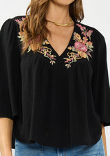 [Color: Black/Dusty Rose] A detail image of a brunette model wearing a black embroidered top with flowy wide sleeves and a v neckline. 
