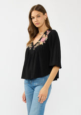 [Color: Black/Dusty Rose] A side facing image of a brunette model wearing a black embroidered top with flowy wide sleeves and a v neckline. 